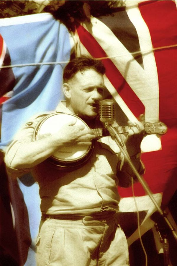 A man wearing WW2 uniform playing a bango in front of a microphone with Union Jack flag in the background